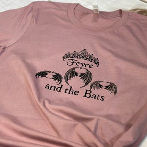 May include: A dusty rose t-shirt with a black graphic design featuring a crown, the text "Feyre and the Bats", and three bat wings.
