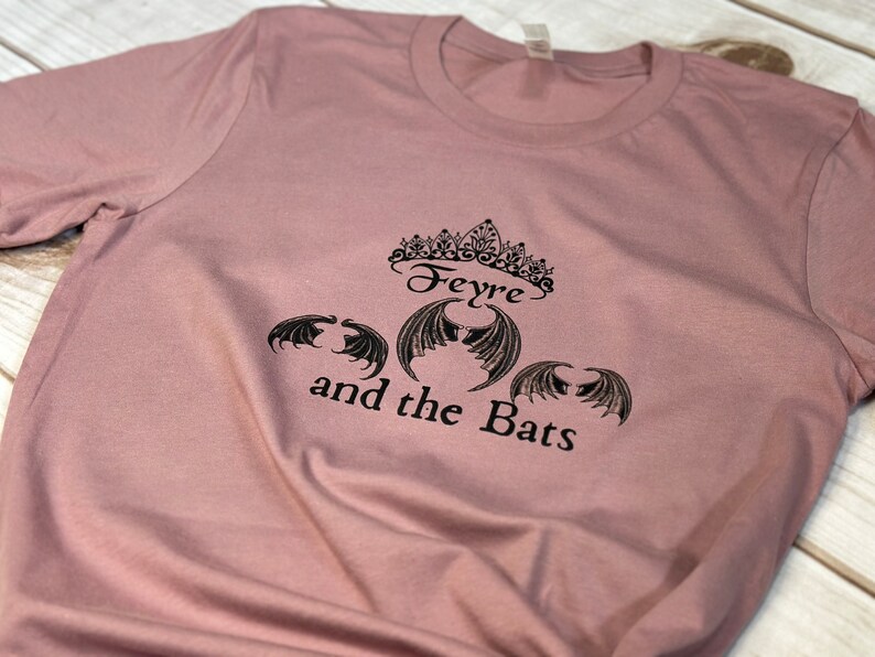 May include: A dusty rose t-shirt with a black graphic design featuring a crown, the text "Feyre and the Bats", and three bat wings.