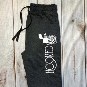 May include: Dark gray sweatpants with a drawstring waist. A white graphic of a lighter, rose, and the word "HOOKED" is printed vertically on the left leg. The pants are laid on a light wood-grain surface.