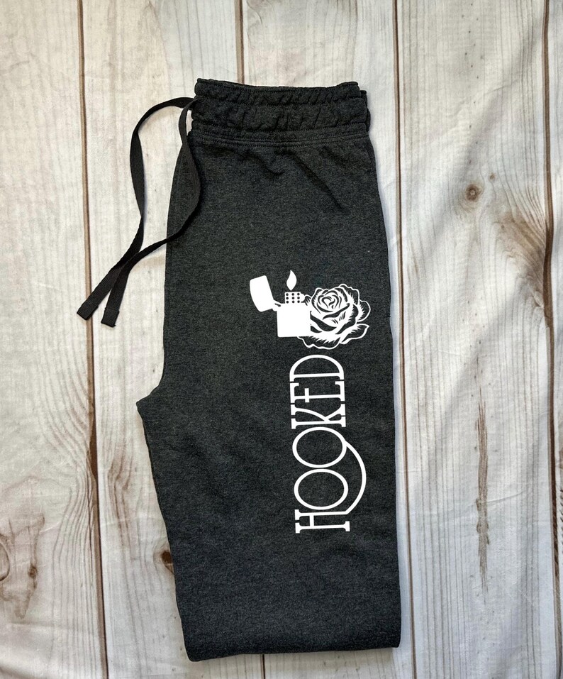 May include: Dark gray sweatpants with a drawstring waist. A white graphic of a lighter, rose, and the word "HOOKED" is printed vertically on the left leg. The pants are laid on a light wood-grain surface.