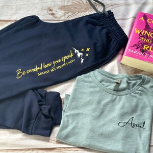 K&ouml;nnte beinhalten: Marineblaue Jogginghose mit dem Text "Be careful how you speak ABOUT MY HIGH LADY" und einer Berg-Grafik. Ein hellblaues T-Shirt mit dem Namen "Azriel" und ein Buch mit dem Titel "A Court of Wings and Ruin" sind ebenfalls im Bild.