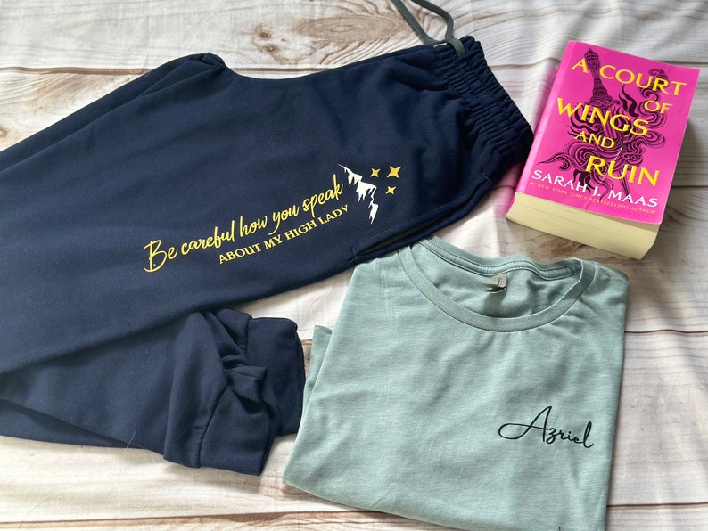 K&ouml;nnte beinhalten: Marineblaue Jogginghose mit dem Text "Be careful how you speak ABOUT MY HIGH LADY" und einer Berg-Grafik. Ein hellblaues T-Shirt mit dem Namen "Azriel" und ein Buch mit dem Titel "A Court of Wings and Ruin" sind ebenfalls im Bild.