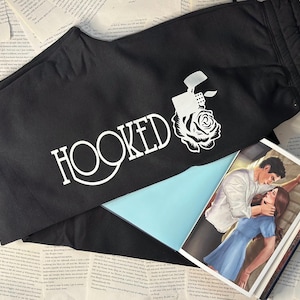 May include: Black sweatpants with the word "HOOKED" in white script, along with a white graphic of a lighter and rose. A book with a romantic illustration is partially visible.
