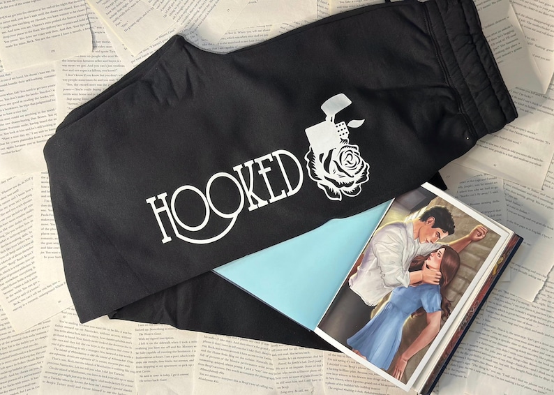 May include: Black sweatpants with the word "HOOKED" in white script, along with a white graphic of a lighter and rose. A book with a romantic illustration is partially visible.