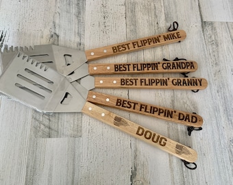 May include: Four wooden-handled grilling spatulas with metal blades. The handles are engraved with the text "Best Flipping'" followed by a name. The names are Mike, Grandpa, Granny, and Dad. The last spatula has the name Doug and a small American flag design.