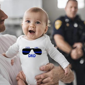 May include: A white baby onesie with a blue line sunglasses graphic and the text "Cool Like Dad". A baby is being held by an adult.