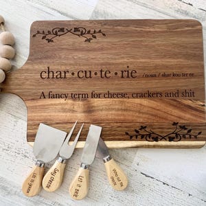 May include: A wooden cutting board with the text "charcuterie /noun/ shar koo ter ee A fancy term for cheese, crackers and shit" engraved on it. The board is accompanied by four small cheese knives with engraved handles that say "Life is GOUDA", "SAY CHEESE", "Let it BRIE", and "Who cut the cheese".