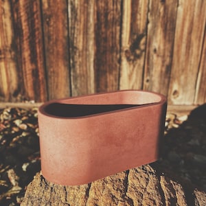 May include: A terracotta-colored oval planter made of concrete. The planter is sitting on a rock.