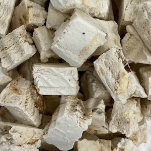 May include: Close-up of a pile of white nougat candy pieces. The candy has a soft, textured appearance with visible inclusions of nuts and a light brown color. The pieces are irregularly shaped and appear freshly made.