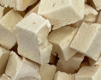 May include: Close-up of a pile of square, off-white marshmallows. The marshmallows have a slightly rough texture and appear to be freshly made. Some pieces are partially cut or broken, revealing a soft interior.