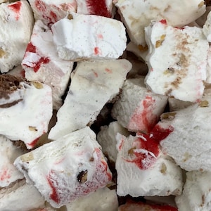 May include: Close-up of a pile of white, textured nougat pieces with red streaks and small nut inclusions. Some pieces have a light brown or chocolate-like appearance. The image is a close-up, showing the texture and details of the confection.