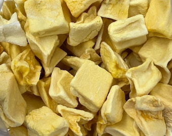 May include: A collection of freeze-dried pineapple chunks, each featuring a pale yellow color and a porous, irregular cubic shape. The pieces have a dry, matte texture with visible fibrous edges and varying shades of light to golden yellow.