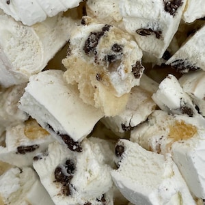 May include: Close-up of a pile of white, irregularly shaped nougat pieces with dark chocolate chips. The nougat has a soft, fluffy texture and a slightly uneven surface, with some pieces showing a light tan color.