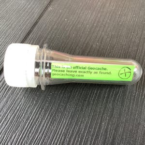 May include: A clear plastic cylindrical geocache container with a white screw-on cap. A bright green label is attached to the side, featuring the black text: This is an official Geocache. Please leave exactly as found. geocaching.com, and a logo.