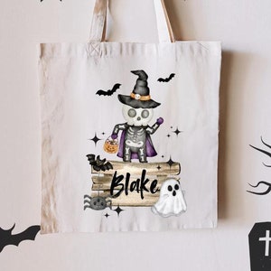 May include: A white canvas tote bag featuring a Halloween-themed design. The design includes a skeleton wearing a witch hat and cape, holding a candy basket. The name "Blake" is printed on a wooden sign. Bats, a spider, and a ghost are also included.