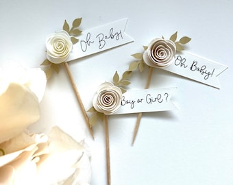 May include: Three white cupcake toppers with gold leaf accents and paper flowers. The toppers have the text "Oh Baby!" and "Boy or Girl?" written on them.