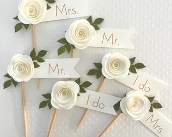 Könnte beinhalten: Sechs dekorative Lebensmittelspieße mit weißen Papierrosen und grünen Papierblättern an Holzspießen. Jeder Spieß enthält ein weißes Banner mit goldenem Text, der entweder Mr., Mrs. oder I do lautet, angeordnet in einem verstreuten Muster.