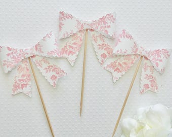 May include: Three pink and white floral patterned paper bow cupcake toppers with scalloped edges on wooden sticks.