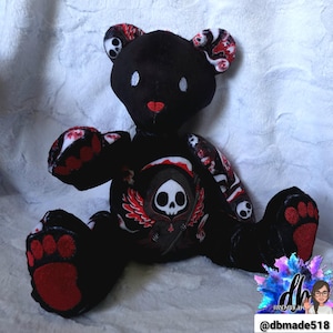 May include: Black plush teddy bear with red and white patterned accents on the ears, paws, and arms. The bear features a grim reaper graphic on its chest with a skull and wings. The bear has white eyes and a red nose.