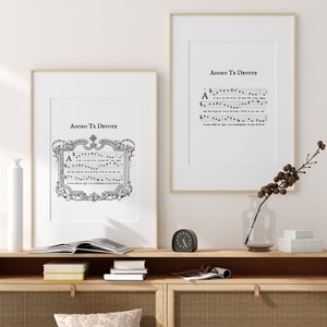 Puede incluir: Dos impresiones enmarcadas de partituras musicales con el t&iacute;tulo "Adoro Te Devote" en texto negro. Las impresiones est&aacute;n colgadas en una pared blanca sobre un estante de madera con un reloj, un jarr&oacute;n y libros.