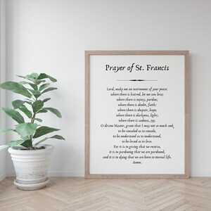 May include: A framed print of the Prayer of St. Francis, a well-known Catholic prayer. The print is on white paper with black text and is framed in a light brown wood frame.