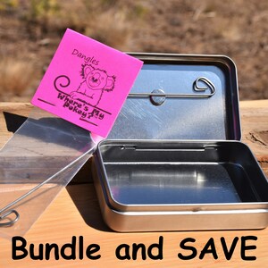 May include: A silver metal tin with a hinged lid and a small metal hook. The tin is open and empty. A pink sticky note with the text "Dangles Where's My Pokie?" and a cartoon koala is attached to the tin. A clear plastic bag with a silver metal hook and a leaf charm is next to the tin.