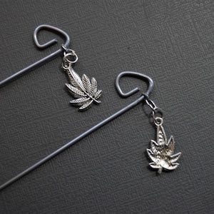 May include: Two silver hair sticks with a silver marijuana leaf charm hanging from each. The hair sticks are on a dark gray textured surface.
