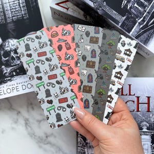 May include: A hand displays five patterned bookmarks. Each bookmark has a different coloured background and a repeating pattern of spooky illustrations, including tombstones, churches, and handcuffs. The bookmarks are in front of books.