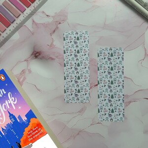 May include: Two white bookmarks featuring a winter-themed pattern. The design includes penguins, hats, mittens, and snowflakes in shades of green, purple, and pink.