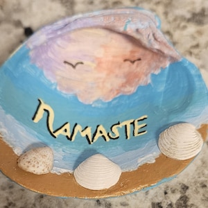 May include: Hand-painted seashell trinket dish with a beach theme. The dish is painted with a blue ocean, a pink and purple sky, and the word "Namaste" in yellow. Three seashells are placed on the dish.