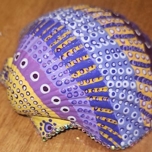 May include: A hand-painted seashell with a vibrant design. The shell features a pattern of yellow, purple, and blue sections, adorned with white dots and lines. The edges are embellished with small, sparkling rhinestones. This decorative item is perfect for coastal decor.