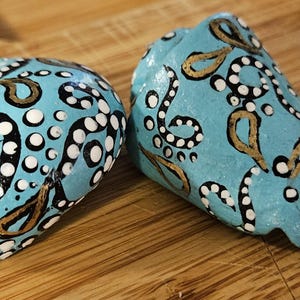 May include: Two hand-painted, turquoise-colored decorative items. Each has a unique design featuring black, white, and gold swirls and dots. The items are displayed on a wooden surface.