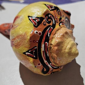 May include: A hand-painted seashell with a vibrant design. The shell features a yellow base with orange and red accents, and black and white decorative patterns. The shell's texture is visible, adding to its unique appeal.