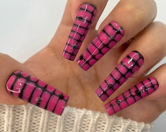 May include: A hand displaying long, coffin-shaped artificial nails. The nails are a vibrant pink with a black web-like design and small, dark embellishments. The nails are set against a white background.