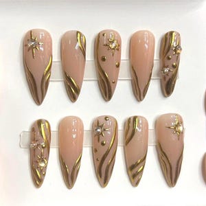 May include: Set of ten nude-colored false nails with gold accents and rhinestones. The nails are long and have a pointed shape.