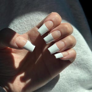 May include: A hand with long, square-shaped white acrylic nails. The nails have a French tip design with a natural base. The hand is resting on a light gray surface, with shadows cast across the hand and surface.