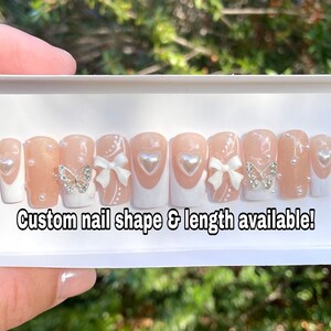 May include: A set of press-on nails with a nude base and white tips. Some nails feature a silver butterfly, white bows, and pearl accents. Text reads: Custom nail shape & length available!