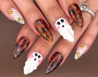 May include: A set of long, almond-shaped fingernails with a Halloween theme. The nails feature a tortoiseshell design with gold flakes, white ghost designs with pink cheeks, and white nails with gold embellishments.