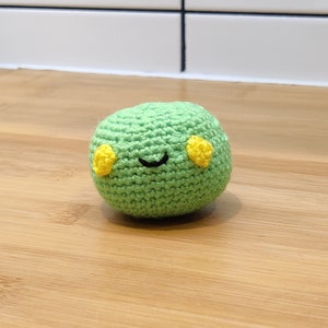 Può includere: Un giocattolo di peluche verde all'uncinetto con occhi gialli e una bocca sorridente nera.