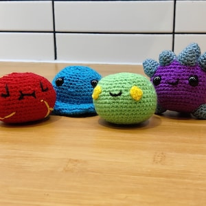 Puede incluir: Cuatro peluches de crochet con diferentes expresiones faciales. Los peluches son rojo, azul, verde y morado.