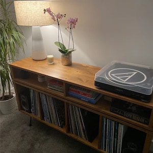 May include: A wooden media console with open shelving units filled with vinyl records. On the top surface sits a white lamp with a textured shade, a small potted pink orchid, a candle, and a black turntable with a clear dust cover.