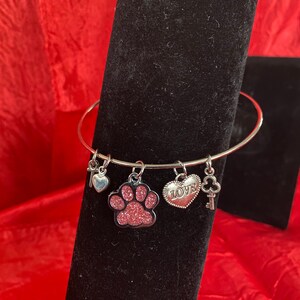 May include: Silver-toned bangle bracelet with multiple charms. Charms include a key, a heart, a pink glitter paw print, a heart with the word "LOVE", and a key-shaped charm. The bracelet is displayed on a black surface with a red background.