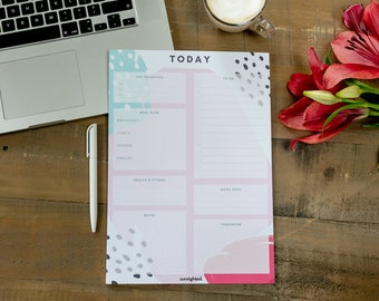 Op de afbeelding: Een roze en blauwe dagelijkse planner met een stippenpatroon. De planner heeft secties voor topprioriteiten, maaltijdplan, te doen, gezondheid en fitness, goede vibes, notities en morgen. De planner ligt op een houten tafel met een laptop, een pen en bloemen.