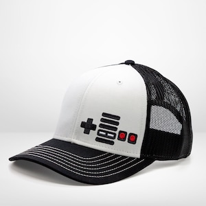 May include: A white and black trucker hat with a black and red video game controller graphic on the front.