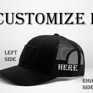 Könnte beinhalten: Schwarze Trucker-Kappe mit Netzrücken. Die Kappe hat eine schwarze gestickte amerikanische Flagge auf der linken Seite und das Wort "HERE" auf der rechten Seite gestickt.