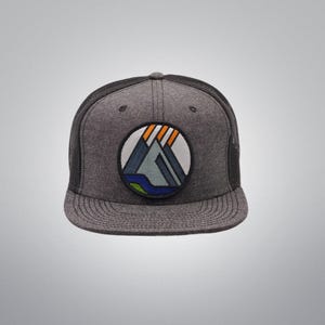 May include: A gray trucker hat with a black mesh back and a curved brim. The hat features a circular patch with a mountain and water design in blue, green, orange, and white. The hat is a casual accessory.