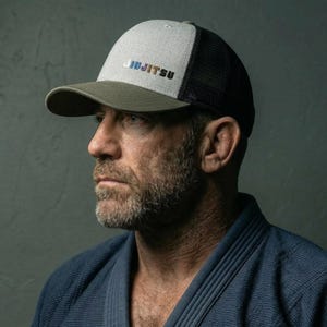 Puede incluir: Gorra de camionero gris y negra con la palabra "JIUJITSU" bordada en azul, verde y amarillo. La gorra tiene una visera verde y una parte trasera de malla.