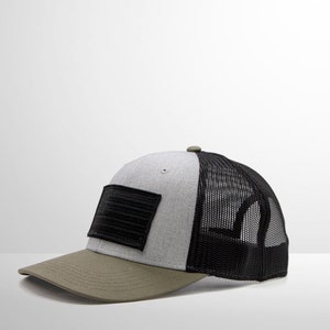 May include: A grey and black trucker hat with a black patch on the front. The hat has a mesh back and a green brim.