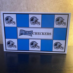 May include: A rectangular game box featuring a blue and white checkerboard pattern. The box displays multiple illustrations of silver football helmets with black panther logos. The center text reads Panthers Checkers in black and blue lettering.