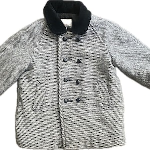 May include: A gray and white tweed coat with a black velvet collar. The coat has a double-breasted design with six black buttons and two front pockets. The sleeves are three-quarter length, and the coat is likely for a child.